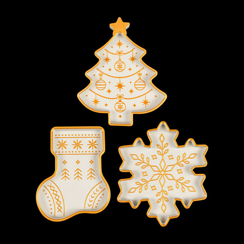 Three white holiday trays with gold trim and patterns: a Christmas tree, a stocking and a snowflake, seen from above on black