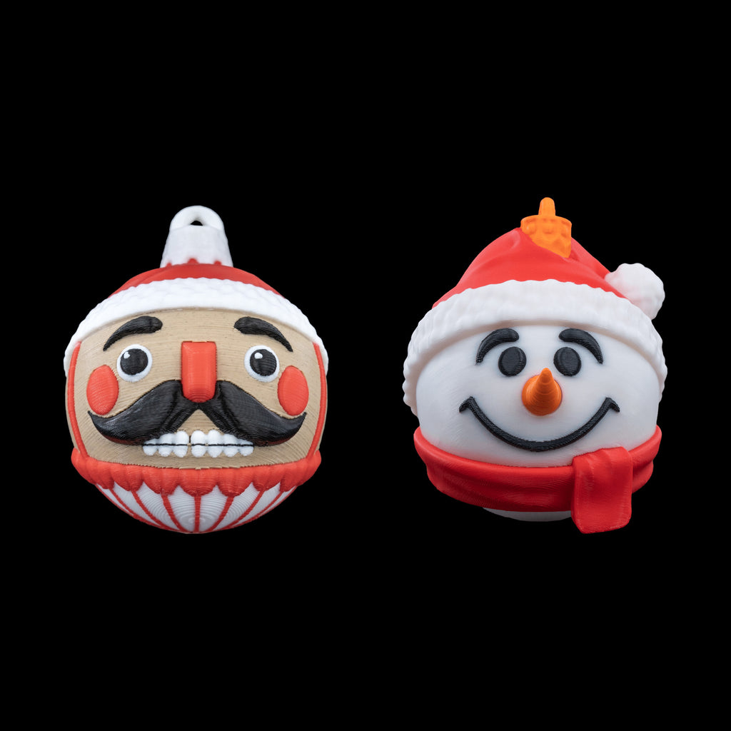 Nutcracker and snowman Christmas ball ornaments side by side, 3D printed in red, white, tan and black, on a black background