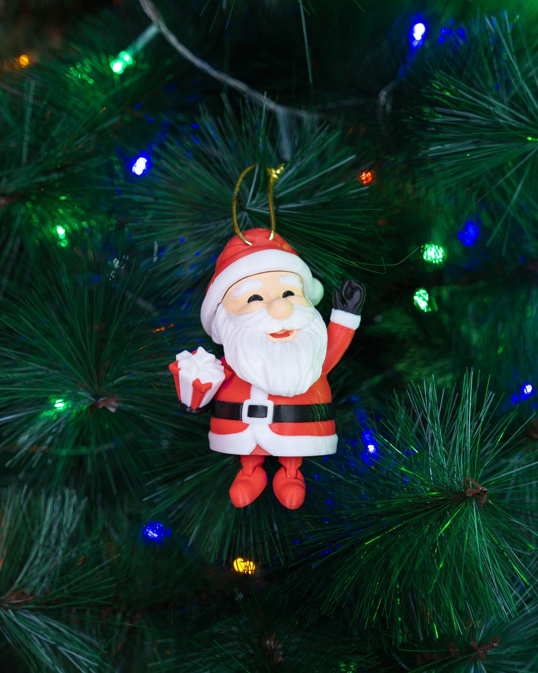 Waving Santa Christmas ornament hanging from its gold cord on a lit Christmas tree, legs dangling